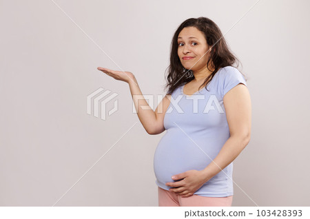 Puzzled perplexed expectant mother, pregnant gravid woman stroking her belly, holding imaginary copy space on her hand palm up, thoughtfully looking at camera isolated on white background. Studio shot 103428393