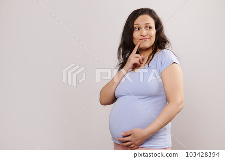 Pensive multi ethnic pregnant woman putting finger near lips, thoughtfully looking aside, gently stroking her belly, isolated on white background with copy space. Maternity leave and pregnancy concept 103428394
