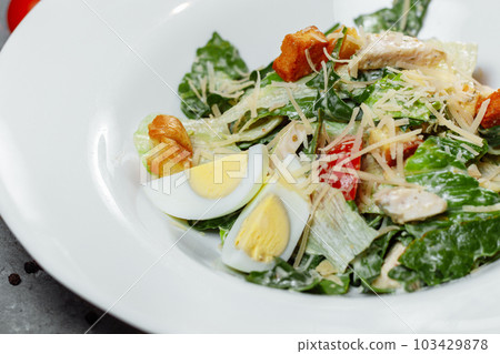 Traditional caesar salad in a white bowl on a old white table with leaves and a sauce boat and parmesan. Selected focus. Traditional caesar salad in a white bowl on a old white table with leaves and a sauce boat and parmesan. Selected focus. 103429878