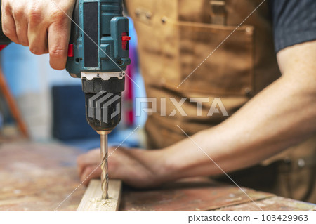 Carpenter drills a hole in wooden plank with an electrical drill 103429963