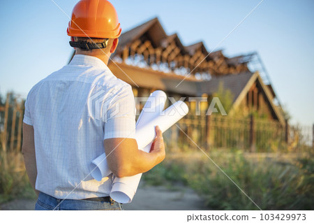 Young engineer in a yellow helmet with blueprints at a construction site 103429973