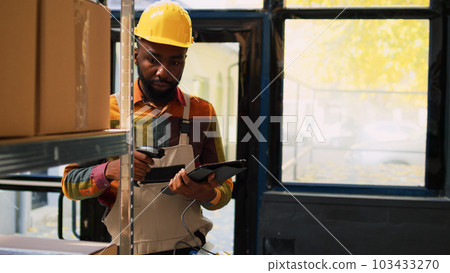 Young employee scanning goods on racks in storage room, working with scanner and tablet for merchandise distribution. Depot worker checking inventory of products before delivery. 103433270