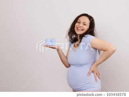 Cheerful multi ethnic positive young pregnant woman with big belly in trimester second of pregnancy, holding cute blue blue knitted baby booties on hand, smiling at camera, isolated white background Cheerful multi ethnic positive young pregnant woman with big belly in trimester second of pregnancy, holding cute blue blue knitted baby booties on hand, smiling at camera, isolated white background 103438578