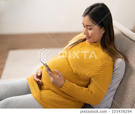 Happy Asian pregnant woman using her phone while relaxing on sofa in her living room 103439294