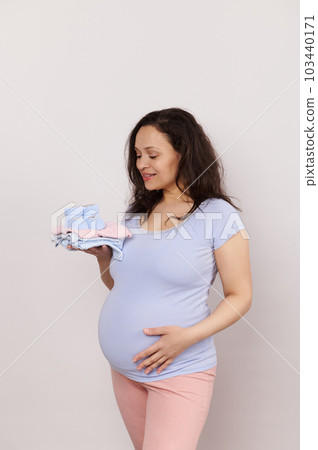 Happy pregnant future mother expecting baby, holds a pile of baby clothes, gently strokes her belly on white isolated background. Gravid female expressing positive emotions of pregnancy and maternity 103440171