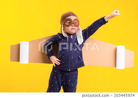 Freedom, girl playing to be airplane pilot, funny little girl with aviator cap and glasses, carries wings made of brown cardboard as an airplane 103445974