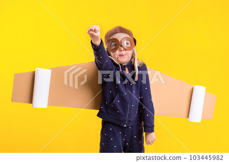 Freedom, girl playing to be airplane pilot, funny little girl with aviator cap and glasses, carries wings made of brown cardboard as an airplane 103445982