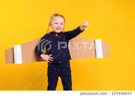 A little child girl is wearing homemade cardboard flying wings, pretending to be a pilot for a craft, imagination or exploration concept. A little child girl is wearing homemade cardboard flying wings, pretending to be a pilot for a craft, imagination or exploration concept. 103446055
