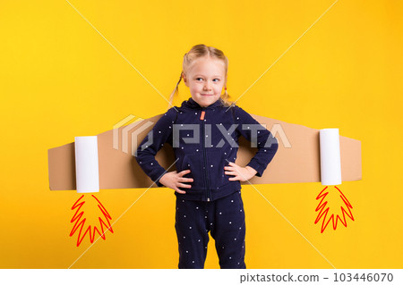 A little child girl is wearing homemade cardboard flying wings, pretending to be a pilot for a craft, imagination or exploration concept. 103446070