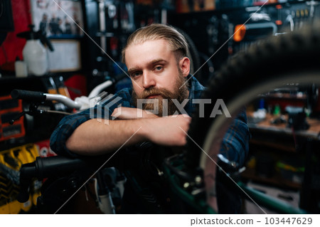 Closeup portrait of cycling repairman standing leaning on bicycle in repair bike workshop with dark interior, serious looking at camera. Concept of professional repair and maintenance of bicycle. 103447629