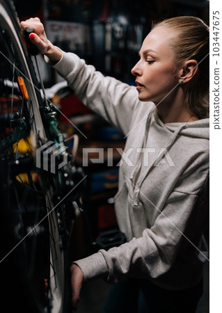 Vertical side view of focused cycling mechanic female repairing and fixing mountain bicycle standing on bike rack in repair workshop with dark interior. Concept of professional bicycle maintenance. Vertical side view of focused cycling mechanic female repairing and fixing mountain bicycle standing on bike rack in repair workshop with dark interior. Concept of professional bicycle maintenance. 103447675