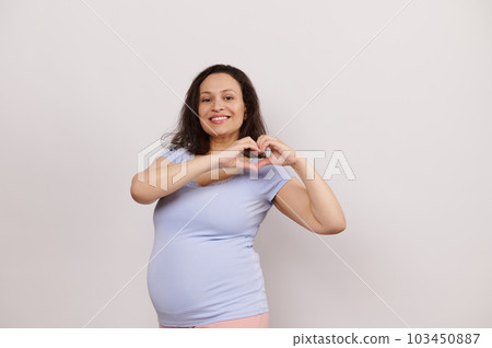 Beautiful pregnant woman smiling at camera, forming a heart shape with fingers, expressing happiness, positive emotions, enjoying her pregnancy and maternity lifestyle, isolated on white background 103450887