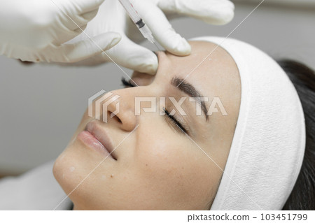 Injection with a syringe with butolin on a woman's face. Care with love: a woman visits a beautician for botulinum therapy, which makes her skin soft, smooth and youthful. Beauty clinic Injection with a syringe with butolin on a woman's face. Care with love: a woman visits a beautician for botulinum therapy, which makes her skin soft, smooth and youthful. Beauty clinic 103451799