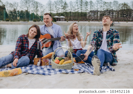 Friends are sitting on a beach blanket, singing and dancing to the guitar, having fun. Cheerful boys and girls enjoying a picnic by the lakeside, drinking beverages, and eating fruits at summer time. 103452116