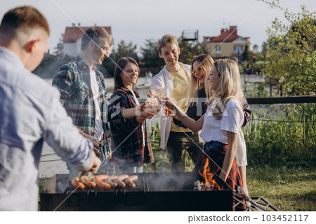 Joyful group of friends is relishing summer vacation with a delightful barbecue. A man grilling sausages over the fire, smoke rises from the meat. Boys and girls are dancing, girl playing the guitar. Joyful group of friends is relishing summer vacation with a delightful barbecue. A man grilling sausages over the fire, smoke rises from the meat. Boys and girls are dancing, girl playing the guitar. 103452117