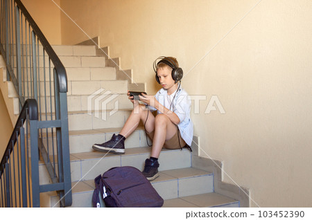 A cute schoolboy is sitting on the stairs with headphones and looking at the phone after school. 103452390