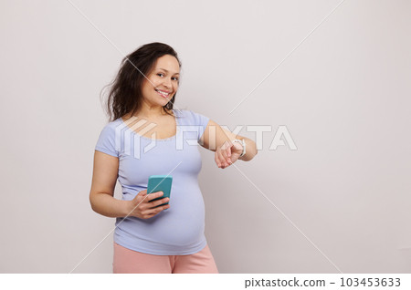 Happy middle aged pregnant woman in 28 week of pregnancy, expecting baby, holding smartphone and checking mobile app on electronic smart wrist watch, smiling looking at camera, over white background 103453633