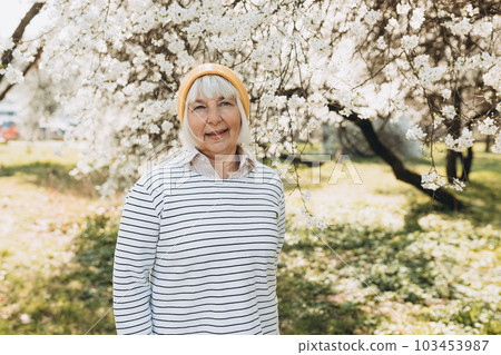 Portrait of elderly happy woman in yellow hat smiling and posing near the blooming cherry tree outdoors, spring day 103453987