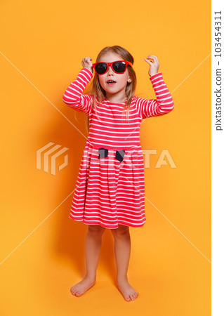 Cute little girl standing in red striped dress and sunglasses on yellow background in the studio. Cute little girl standing in red striped dress and sunglasses on yellow background in the studio. 103454311