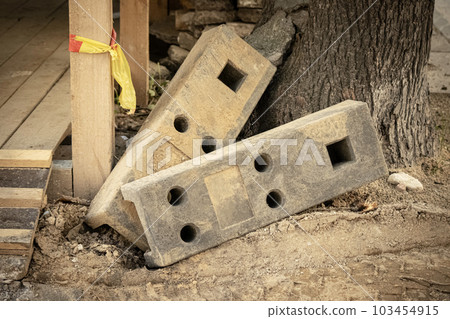 Heavy concrete weight blocks with various holes for temporary construction fence lying near wooden construction and a tree trunk on sand 103454915