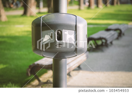 Plastic bag dispenser for dog poo in park with visible benches and green grass lawns on the background on sunny day 103454933