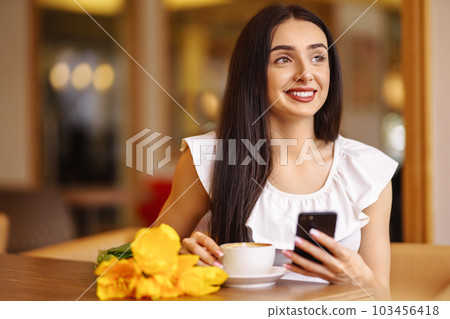 Beautiful Woman With Spring Tulips Flowers Bouquet At Cafe. Woman Drinking Coffee And Using Phone. 103456418