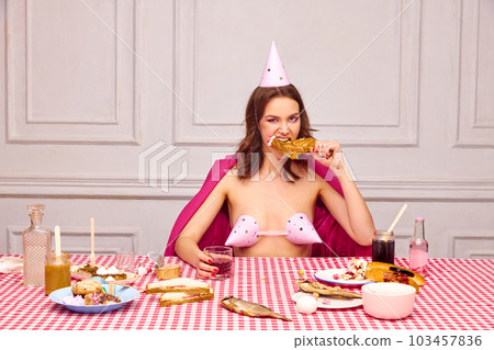 Portrait of beautiful young girl with bright makeup, sitting at table, celebrating her birthday, eating chicken and drinking 103457836