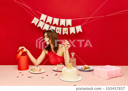 Young girl sitting at table, drinking alcohol, looking at presents and celebrating her birthday against red background Young girl sitting at table, drinking alcohol, looking at presents and celebrating her birthday against red background 103457857