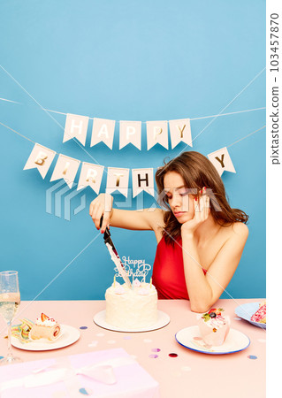 Beautiful young girl sitting at table, celebrating birthday, cutting cake with knife against blue background 103457870