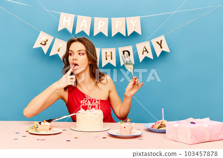Beautiful young girl celebrating birthday, eating cake, drinking champagne against blue background. Pretty female model with bright makeup 103457878