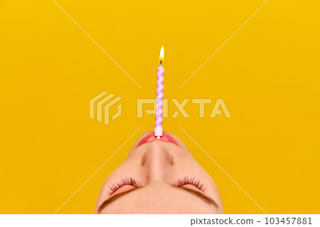 Cropped part of female face with burning candle against vivid yellow background. Making birthday wishes 103457881