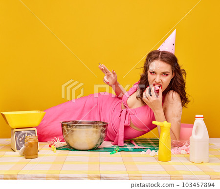 Young birthday girl in pink dress an bright makeup, lying on cooking table, smoking and eating candy against vivid yellow background Young birthday girl in pink dress an bright makeup, lying on cooking table, smoking and eating candy against vivid yellow background 103457894