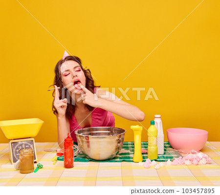 Young beautiful girl with bright makeup eating cake, cooking and smoking against vivid yellow background. Birthday preparation 103457895
