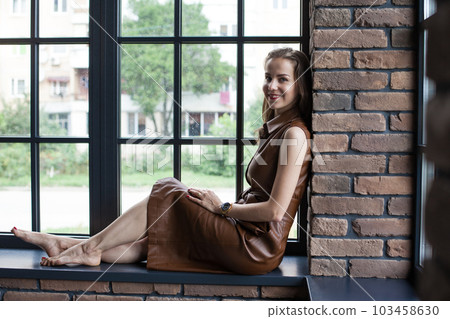 Beautiful young woman in brown leather dress sits on the windowsill barefoot 103458630