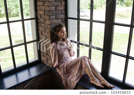 Beautiful young woman in pink dress sits on the windowsill barefoot Beautiful young woman in pink dress sits on the windowsill barefoot 103458655