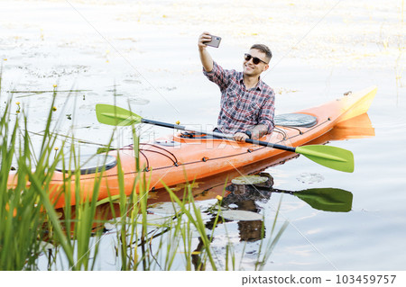 Young Caucasian man sitting in a kayak and taking a selfie on a mobile phone. The concept of water entertainment. 103459757