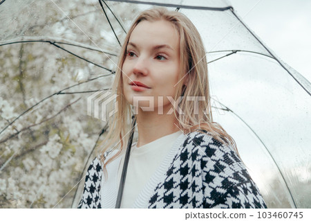 Woman in Plaid Jacket Under a Transparent Umbrella Walks in a Garden with Blooming Trees in Spring Woman in Plaid Jacket Under a Transparent Umbrella Walks in a Garden with Blooming Trees in Spring 103460745