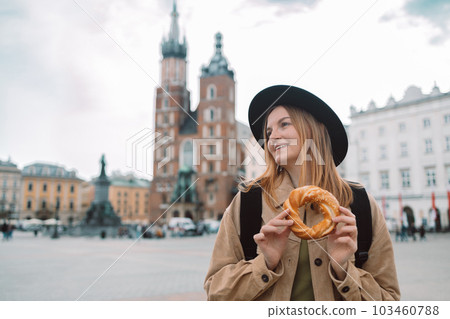 Attractive young female tourist with stylish clothes and a boho hat eating bagel obwarzanek traditional polish cuisine snack waling on Market square in Krakow. Traveling Europe Attractive young female tourist with stylish clothes and a boho hat eating bagel obwarzanek traditional polish cuisine snack waling on Market square in Krakow. Traveling Europe 103460788