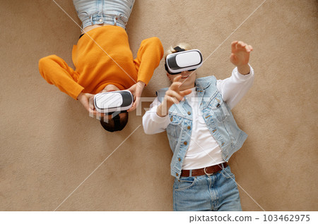 Attractive lesbian woman couple using vr goggles headset lying on floor Attractive lesbian woman couple using vr goggles headset lying on floor 103462975