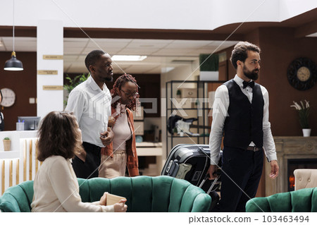 Bellboy taking care of guests luggage, carrying suitcases to room after registration process at reception counter. Male porter assisting tourists to check in and move trolley bags. 103463494