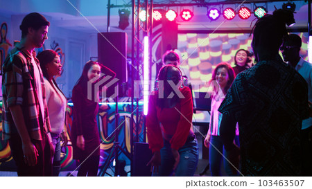 Group of friends partying together on dance floor, listening to live music on stage at nightclub. Young men and women feeling happy at discotheque, dancing and jumping. Handheld shot. 103463507