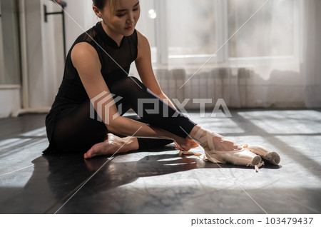 An Asian ballerina sits on the floor and puts on pointe shoes. An Asian ballerina sits on the floor and puts on pointe shoes. 103479437