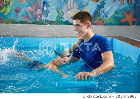 Trainer teaching little boy how to swim in indoor pool with pool floating board 103479968