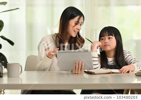 Little cute girl feeling tired and bored while doing her homework or remote studying with mother at home 103480203