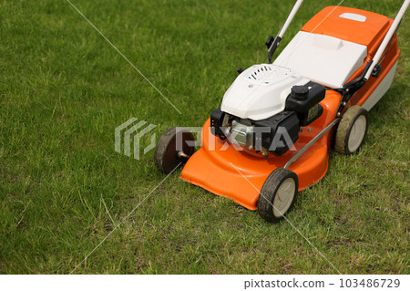 Top view of modern orange-grey electric lawn mower cutting bright lush green grass. Gardening work tools. Rotary lawn mower machine on lawn. Professional lawn care service. Place for text Top view of modern orange-grey electric lawn mower cutting bright lush green grass. Gardening work tools. Rotary lawn mower machine on lawn. Professional lawn care service. Place for text 103486729