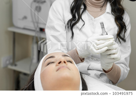 The beautician is preparing to carry out fractional mesotherapy, introducing nutrients to improve complexion, narrow pores and reduce pigmentation. A young woman in a beauty clinic on the procedure. The beautician is preparing to carry out fractional mesotherapy, introducing nutrients to improve complexion, narrow pores and reduce pigmentation. A young woman in a beauty clinic on the procedure. 103491975
