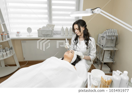 Photo of cosmetic procedure: the picture shows a cosmetologist who skillfully conducts fractional mesotherapy to improve the beauty and health of the skin of a young woman in a beauty clinic Photo of cosmetic procedure: the picture shows a cosmetologist who skillfully conducts fractional mesotherapy to improve the beauty and health of the skin of a young woman in a beauty clinic 103491977