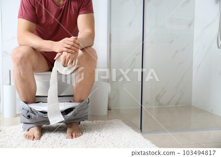 Man with paper roll sitting on toilet bowl in bathroom, closeup 103494817