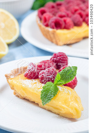 Piece of lemon tart on white plate with fresh raspberry, icing sugar, mint, vertical closeup Piece of lemon tart on white plate with fresh raspberry, icing sugar, mint, vertical closeup 103498420