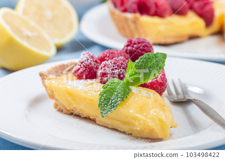 Piece of lemon tart on white plate, garnished with fresh raspberry, icing sugar, mint, horizontal 103498422
